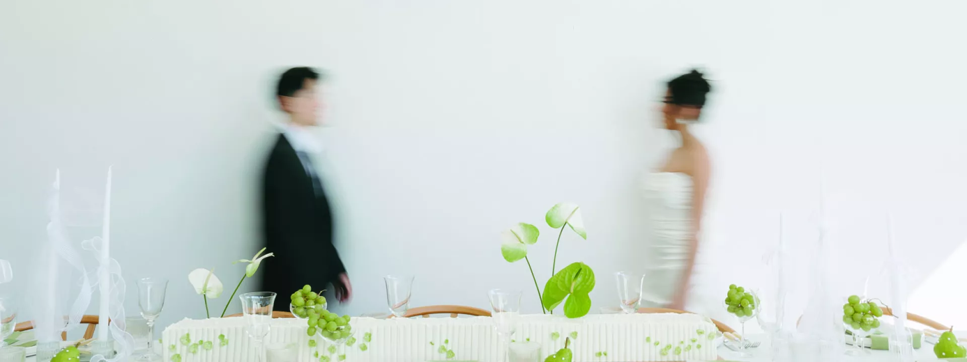 Modern wedding tablescape featuring minimalist white decor, green accents, fresh greenery, and sleek tableware, with a blurred bride and groom walking in the background.