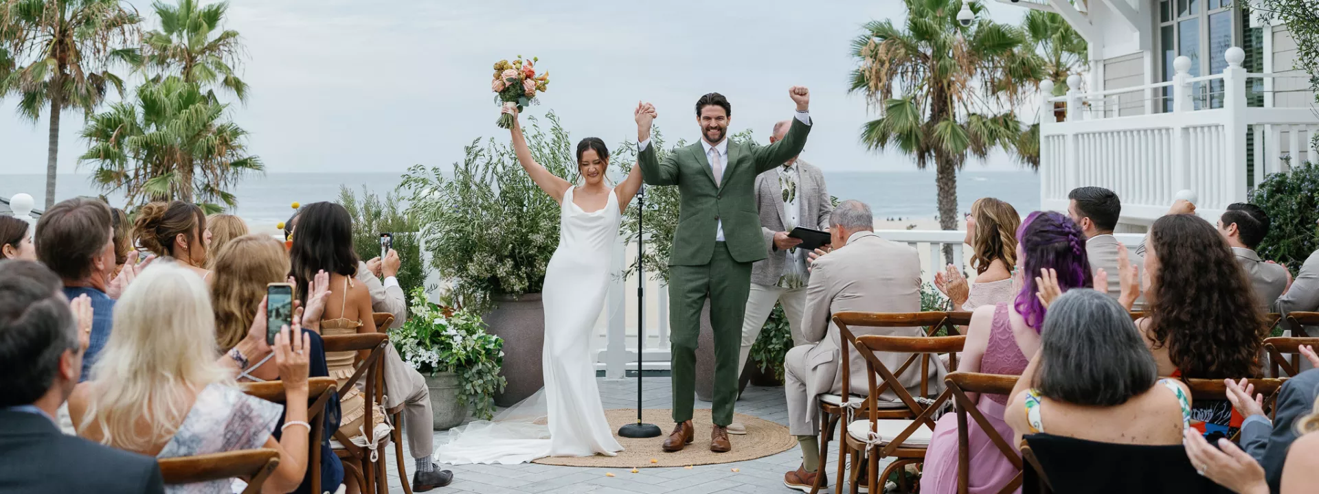 Bride in sleek slip gown and groom in green suit at altar on patio overlooking Santa Monica beach