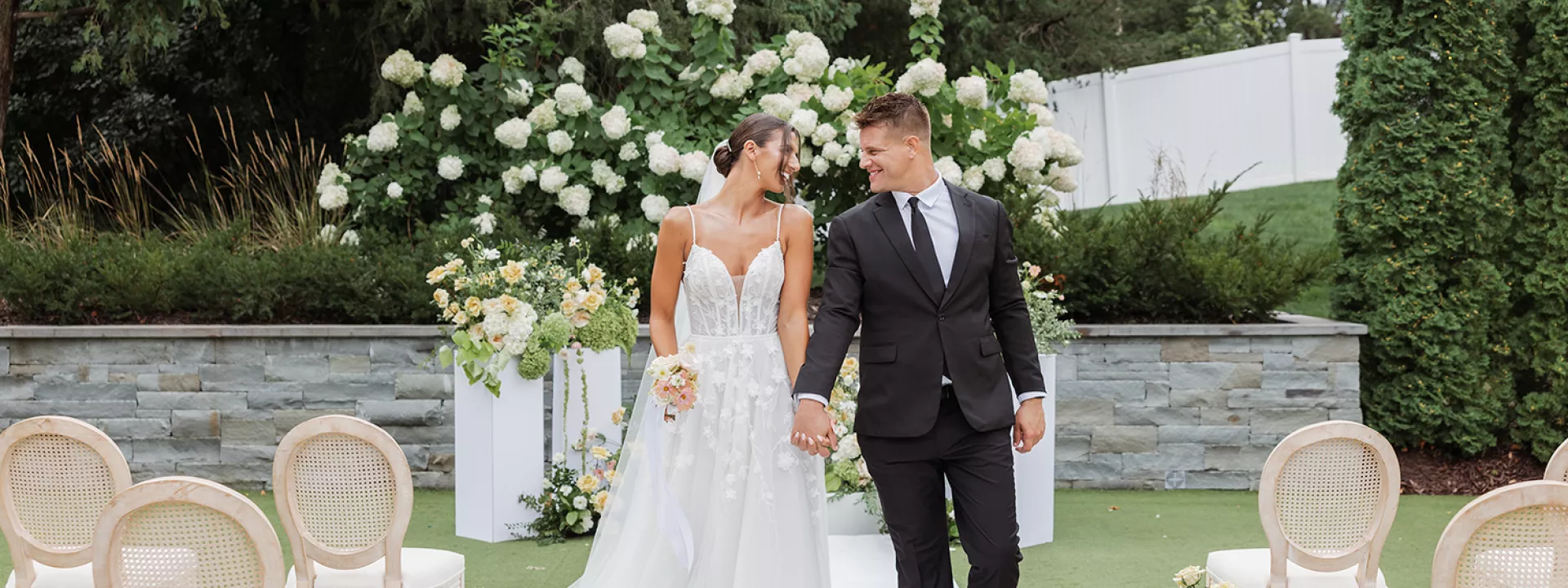 Garden wedding ceremony inspiration featuring a couple walking down a white aisle lined with floral arrangements and rattan chairs during a summer styled shoot at The Hutton House in Minnesota
