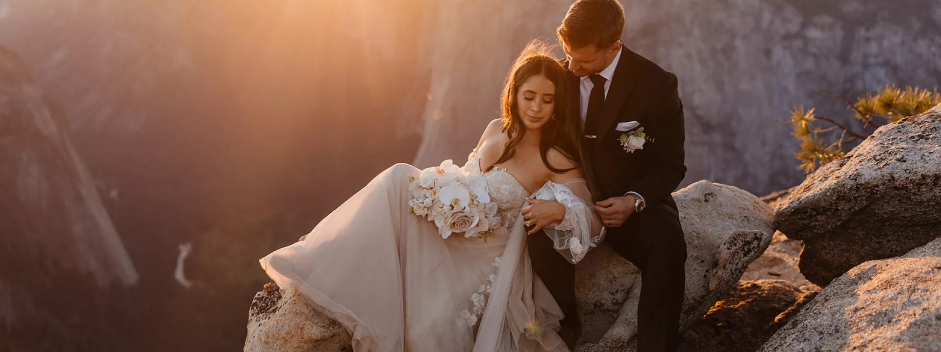 Bride and groom sitting together on a rock at Glacier Point in Yosemite National Park, with the sun setting behind them, creating a golden glow.