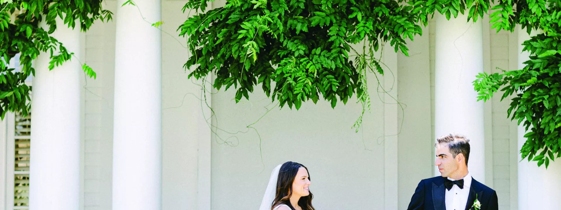 Bride and groom hold hands outside white venue with lush greenery
