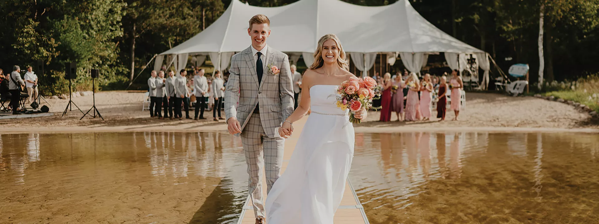 Bride and groom stand holding hands at the end of a dock with wedding party and tented reception behind them in lakeside wedding day