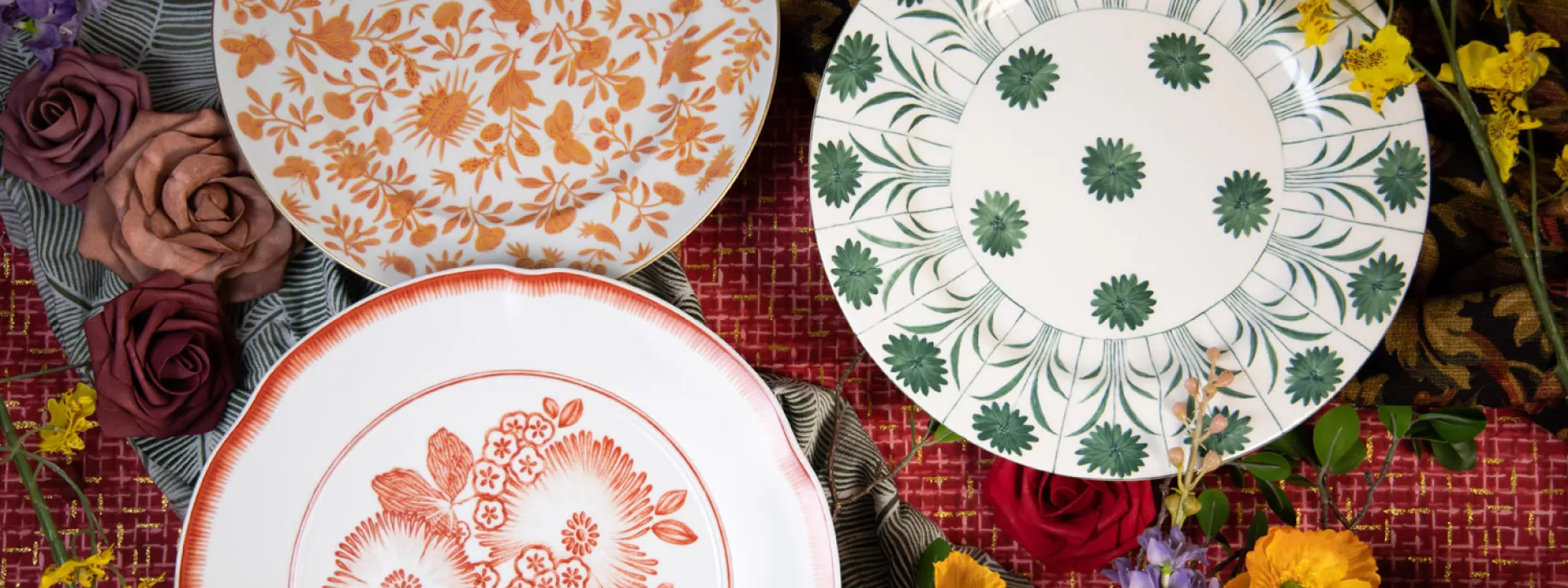 Three designer plates from Maison de Carine—orange Sacred Bird and Butterfly, green Topiary, and coral Oscar’s Coral—styled with roses, poppies, and patterned linens