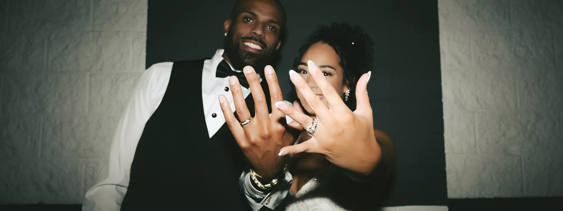 Bride and groom flaunt their rings, holding their hands up as they stand against a black and white checkered background