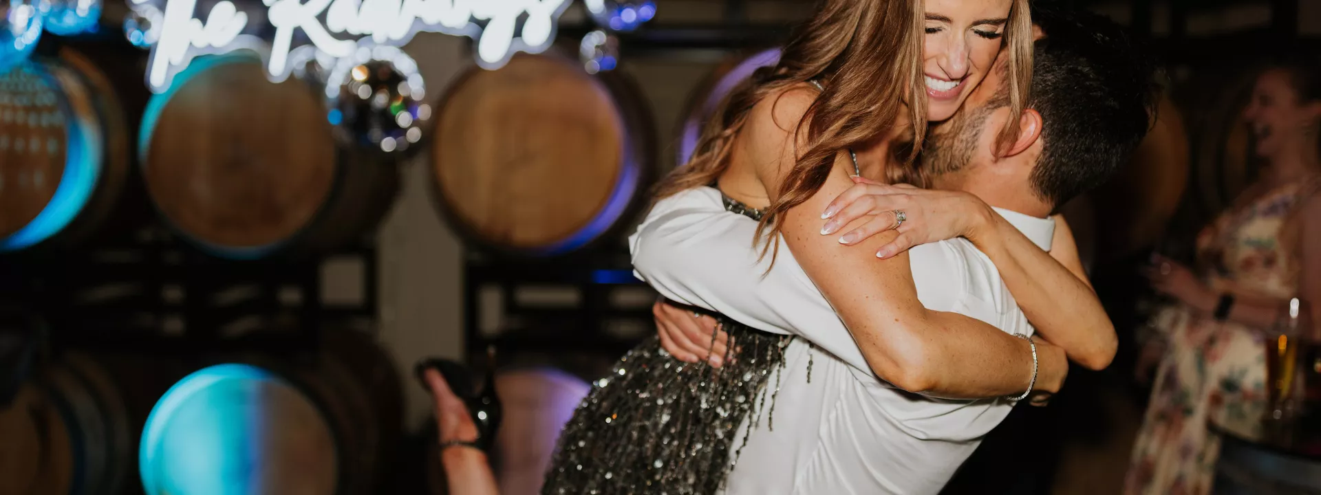 Bride and groom happily embrace in their festive after party outfits in front of a neon sign and wine barrels at JM Cellars