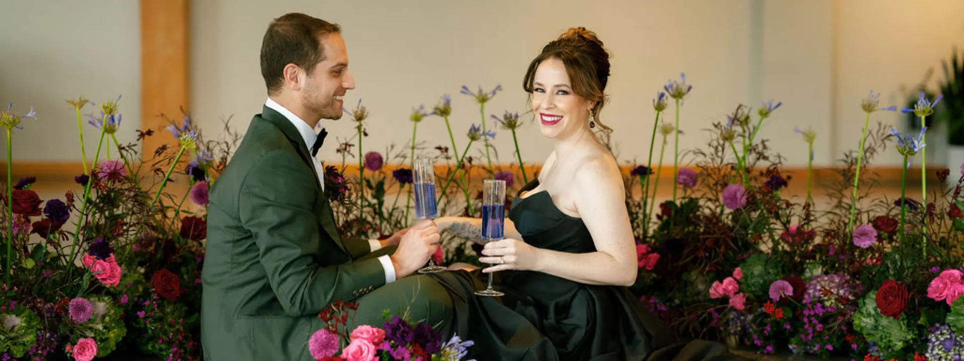 Bride and groom sitting on the floor surrounded by bold winter florals, sharing a drink during their colorful indoor wedding celebration