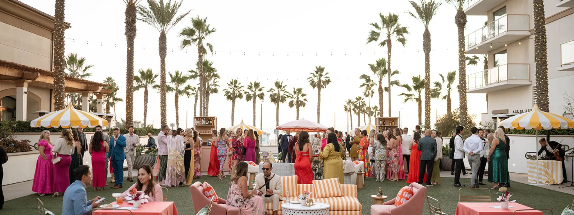 Guests at the Curate Gala 2024 enjoy an elegant outdoor cocktail hour with vibrant lounge furniture, striped umbrellas, and palm tree views at the Waterfront Beach Resort in Huntington Beach