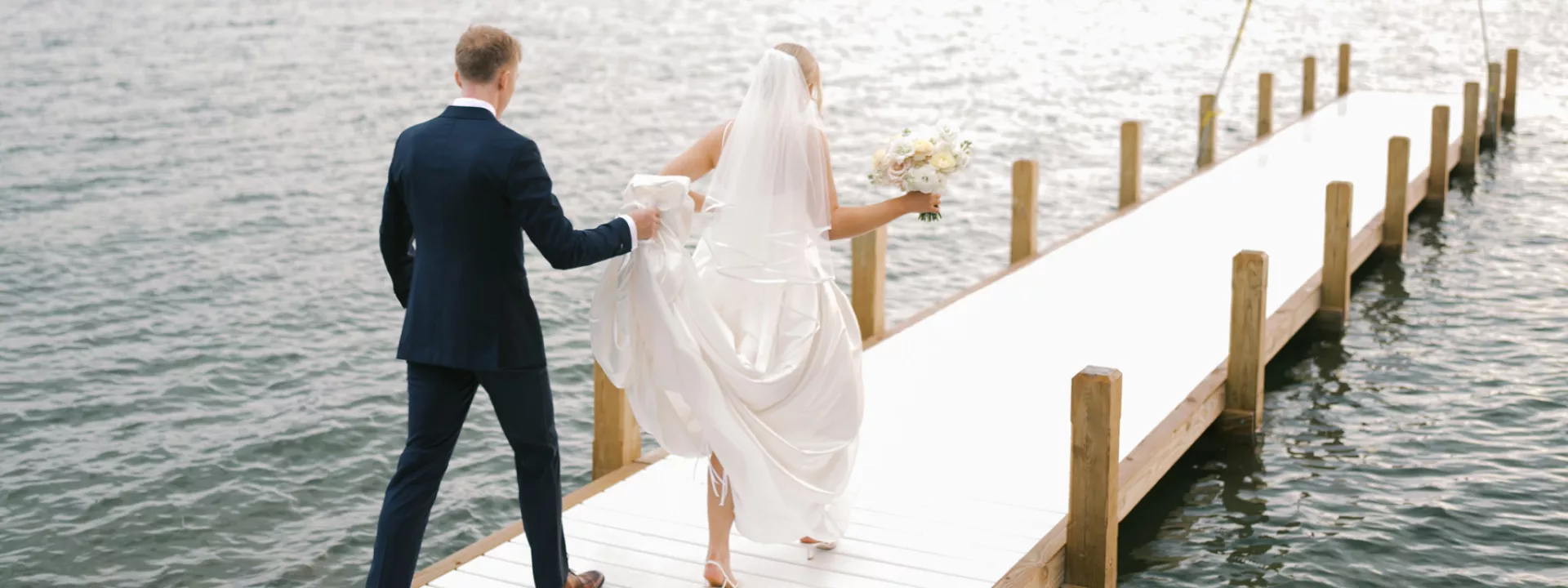 Grace and Lucas walk out on a dock of a lake in their wedding day attire