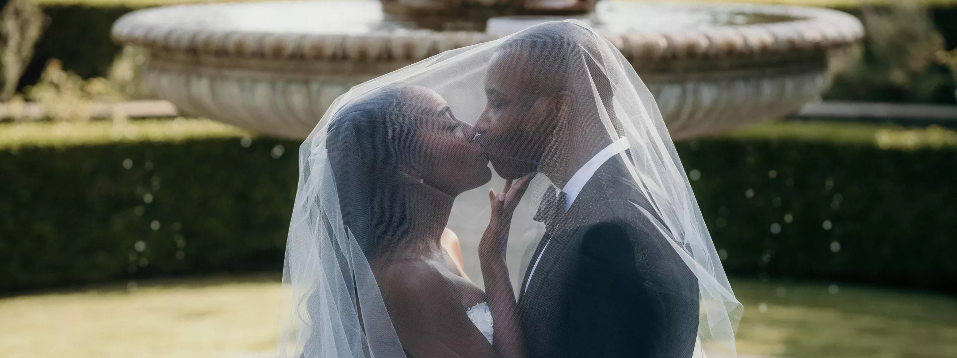 Bride and groom kiss under bride's veil in front of a fountain at Greystone Mansion