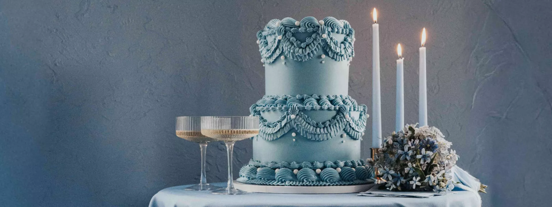 Dusty blue styled scene featuring two-tiered lambeth cake, candles and two glasses of champagne on silky tablecloths