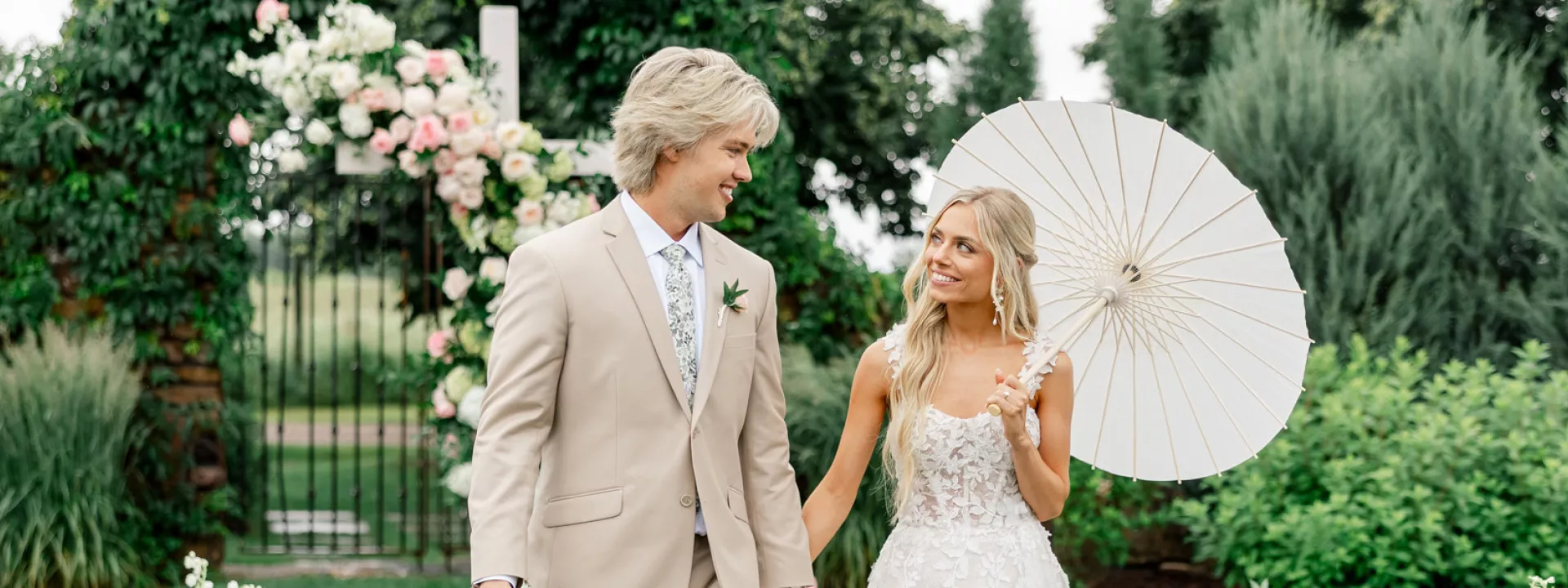 Bride and groom pose in the middle of their outdoor aisle lined by pale pink blooms at Redeemed Farm, bride has a paper umbrella in her hand