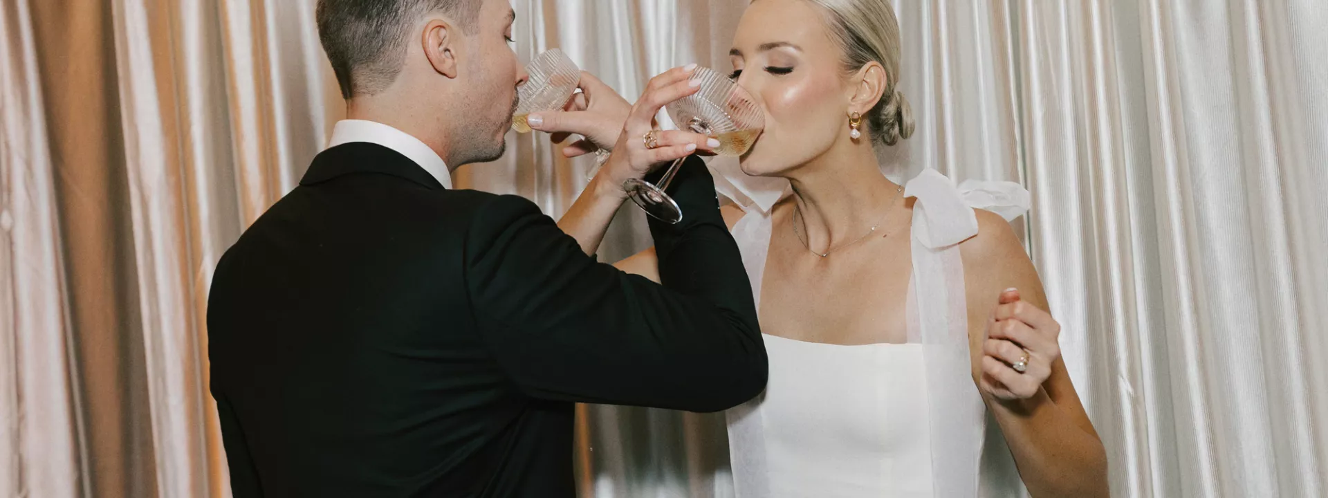 Bride and groom intertwine arms, sipping champagne against a draped backdrop 