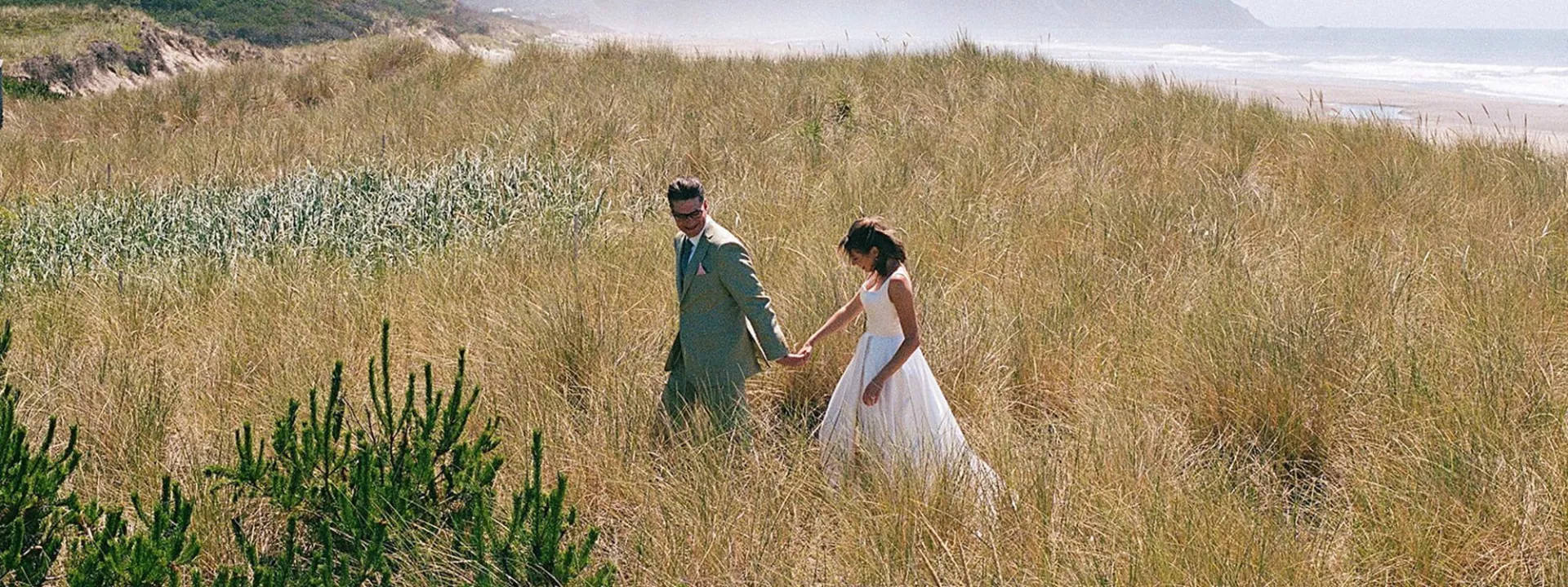 Bride and groom walk hand in hand through oceanside field in coastal wedding day