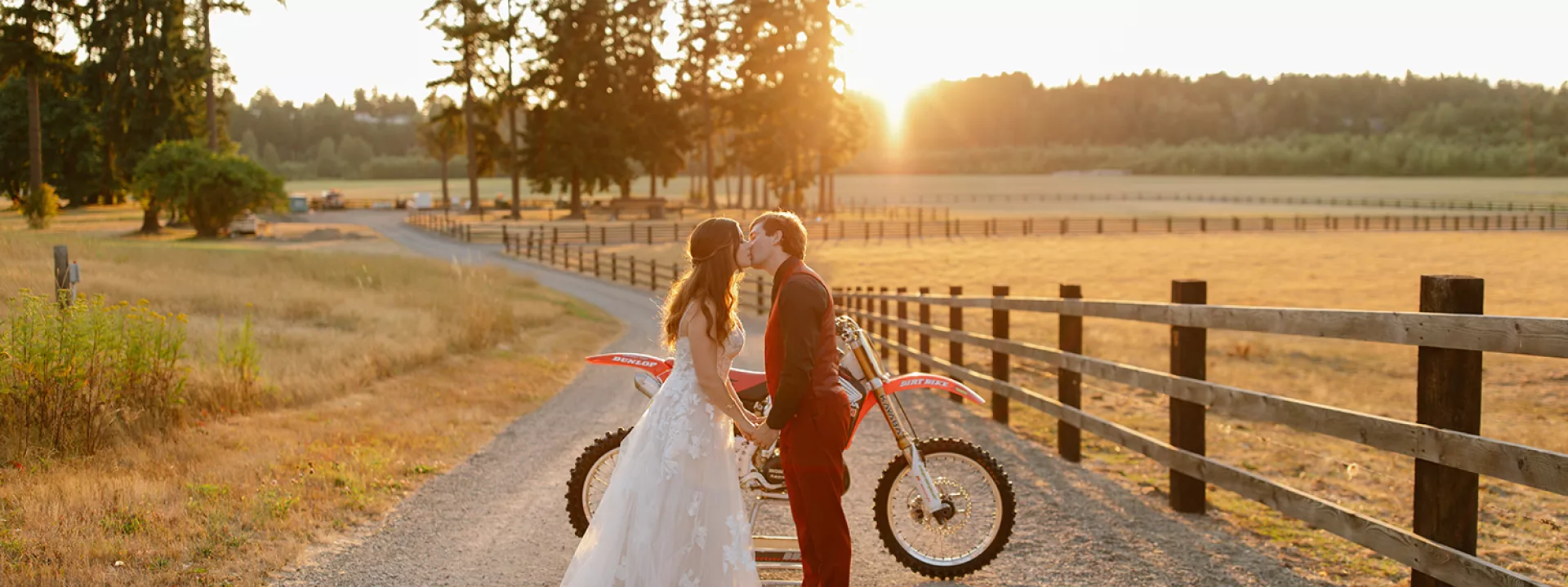 Bride in elegant gown and groom in red suit kiss at sunset with dirt bike in background