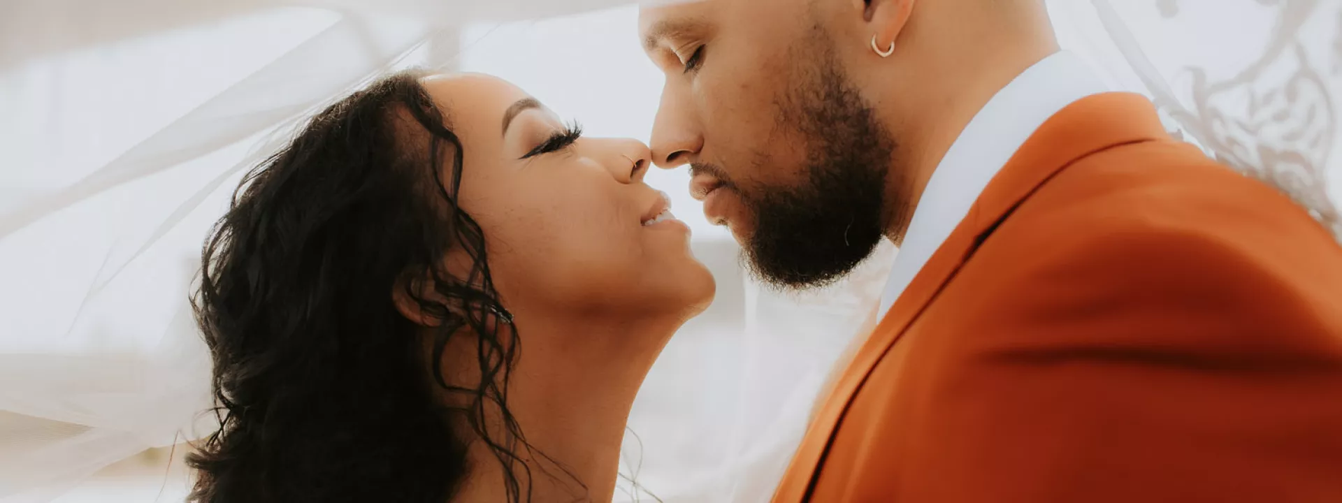 Bride and groom share an intimate moment under the bride’s veil on their fall wedding day, with the groom in a burnt orange suit and the bride in a white lace gown.