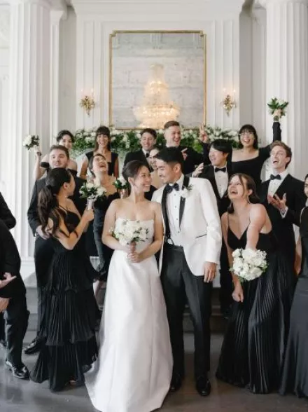 Wedding party featuring an elegant, classic bride and groom surrounded by bridesmaids in black dresses and groomsmen in tuxedos holding white floral bouquets by Poppy Flowers
