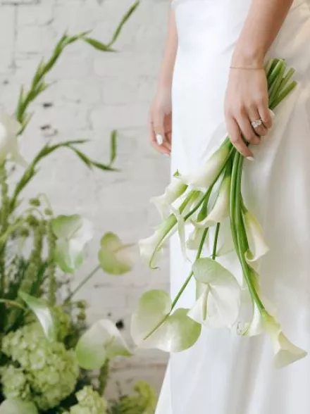 Close-up of a bride holding a minimalist bouquet of white calla lilies and anthurium leaves, showcasing her elegant diamond engagement ring