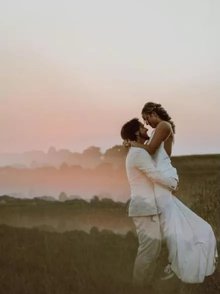 Groom lifts Bride in embrace in a field at sunset