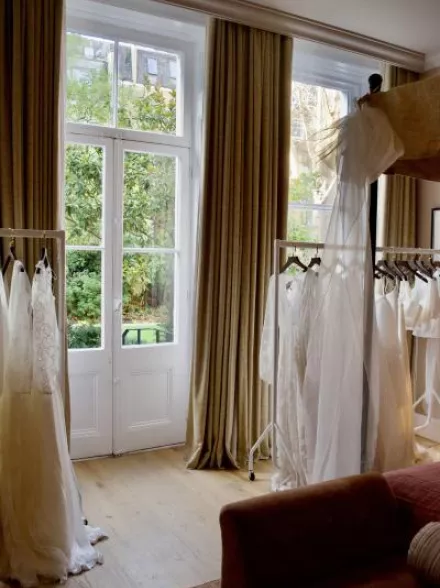 Bridal gowns from E&W Couture displayed on garment racks inside a London suite during the New Archive collection launch, with natural light and garden views in the background.