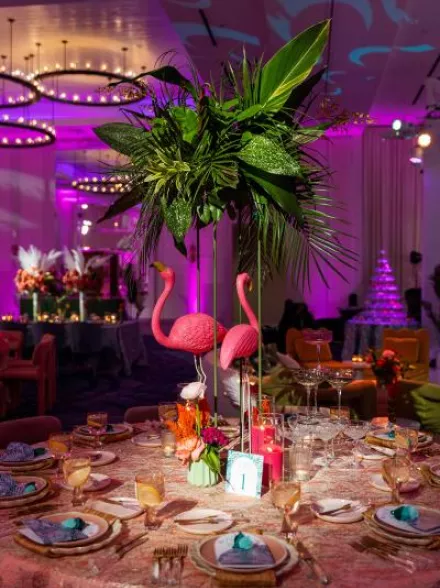 A vibrant dining table at the Curate Gala 2024 features pink flamingo centerpieces, lush tropical greenery, gold flatware, and teal napkins, embodying the Palm Royale theme in the ballroom decor