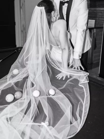 Bride and groom lean against the pool table with pool balls scattered throughout the bride's veil, in black and white