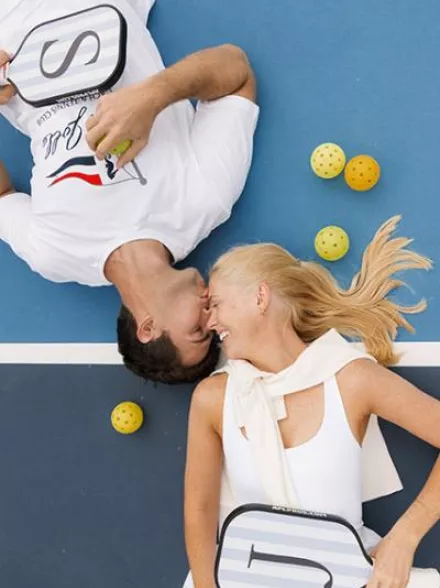 Jennifer and Sean lay on the pickleball court facing each other surrounded by pickleballs